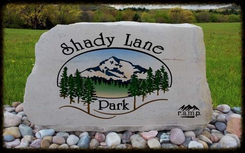 Shady Lane Park sign on a stone slab with mountain, trees, and grass background