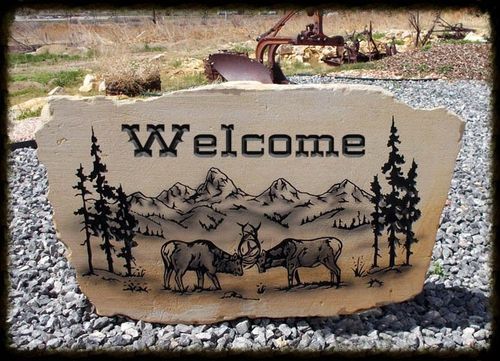 Welcome sign engraved on a stone slab with mountains, trees, and two elk in a rocky outdoor setting