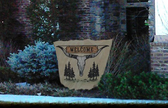 Welcome sign with trees in a landscaped yard beside a brick house.