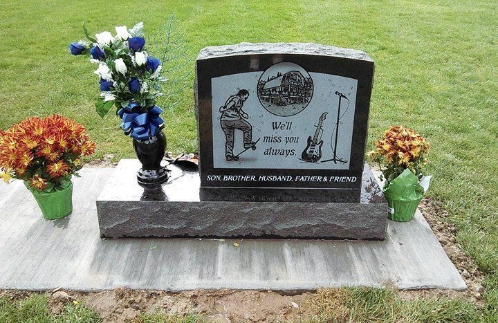 Memorial gravestone on a lawn, decorated with blue and white flowers and small potted plants.