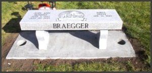 White stone memorial bench engraved “BRAEGGER” on a concrete base in a grassy setting