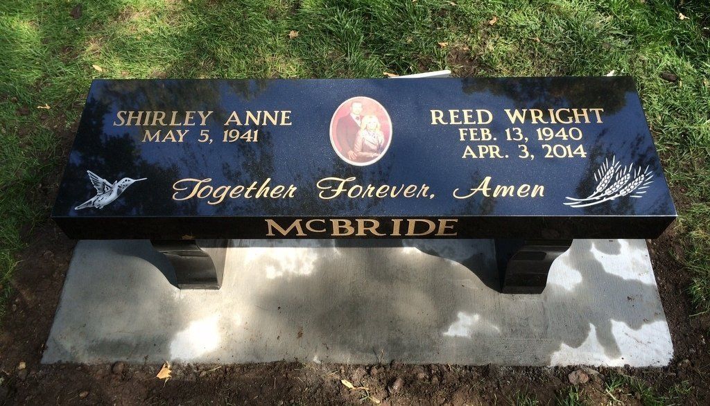Black memorial bench with engraved names and dates set on a stone base in grass and sunlight