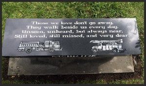 Gray memorial plaque with white inscription and two engraved trucks, set on grass.