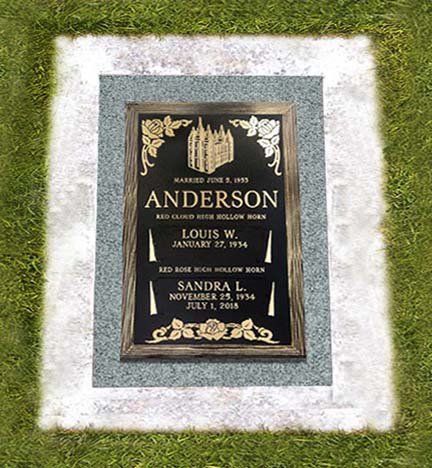 Grave marker with black-and-gold plaque set in a white stone border on grass.