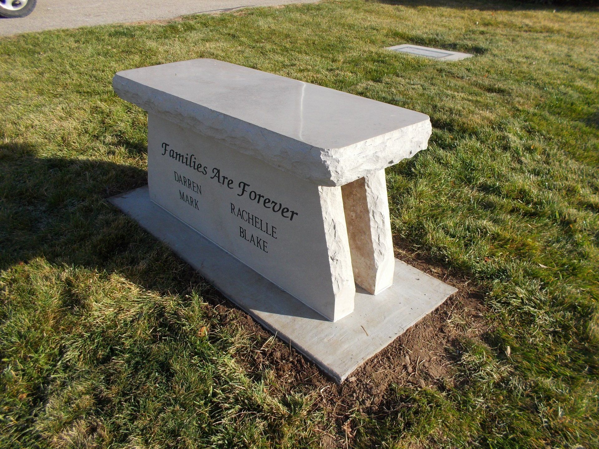 A small stone bench memorial outdoors on grass, with engraved text on the side.