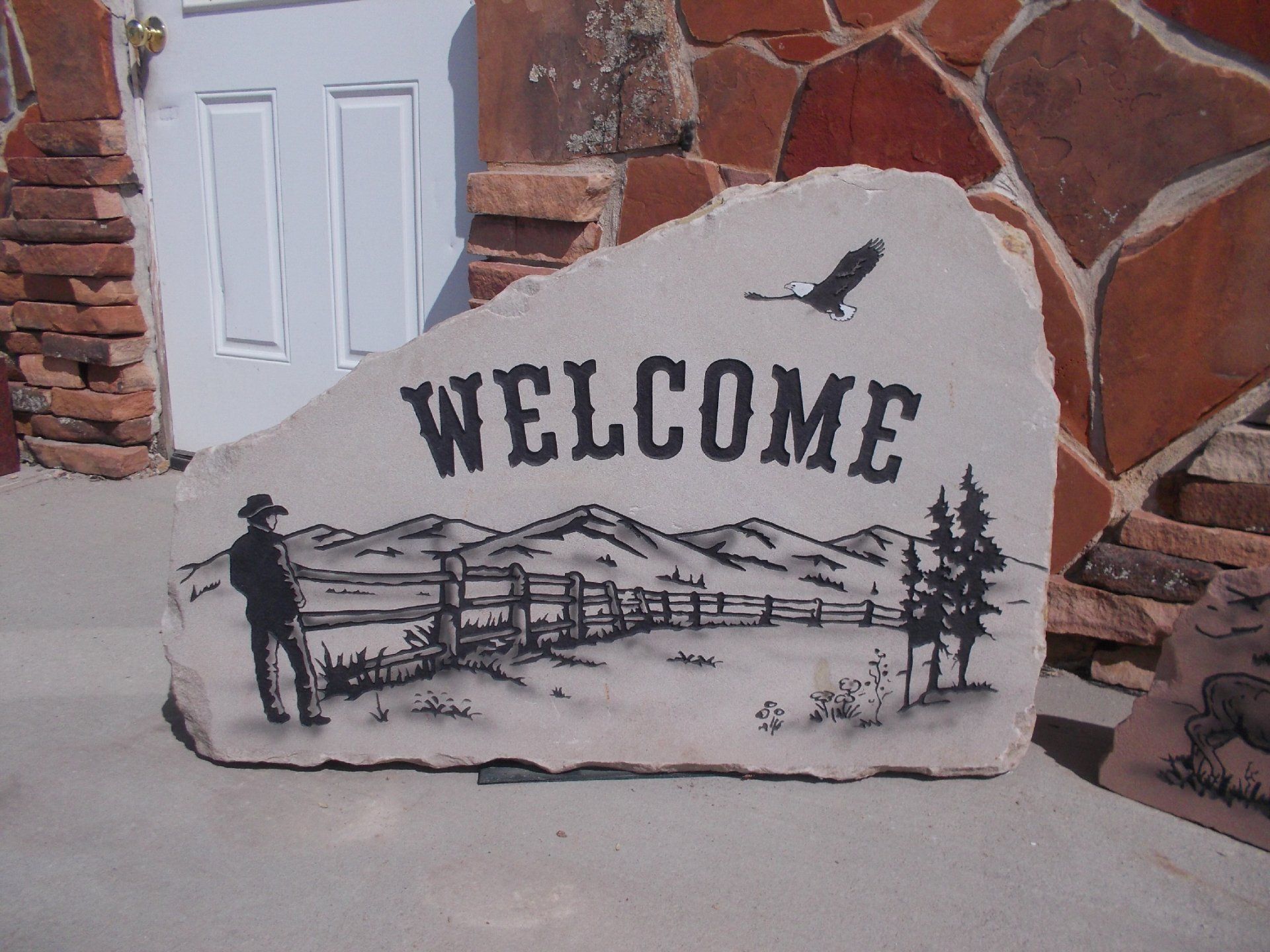 Stone welcome sign with cowboy and mountain scene in black on a white rock.