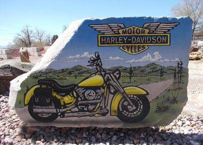 Harley-Davidson sign painted on a large rock with a yellow motorcycle illustration outdoors