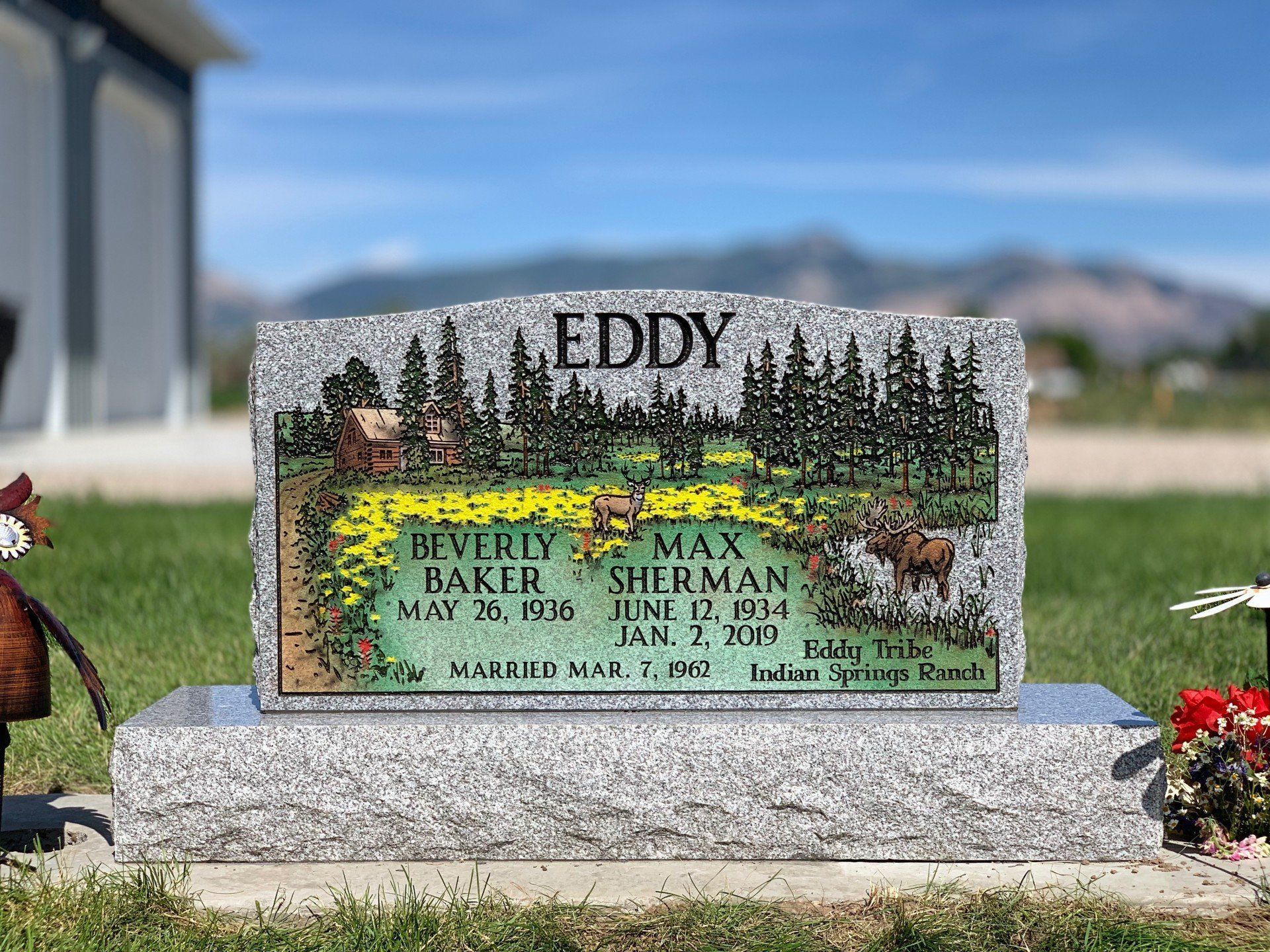 Granite memorial plaque with “EDDY” and names, set in a cemetery with trees and mountains behind.