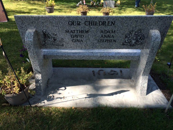 Stone memorial bench in a grassy park, engraved “OUR CHILDREN” with several names.