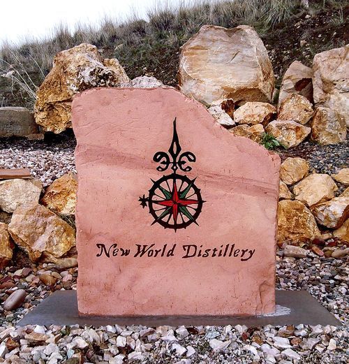 Pink stone sign for New World Distillery with black logo, set outdoors on rocks and grass.