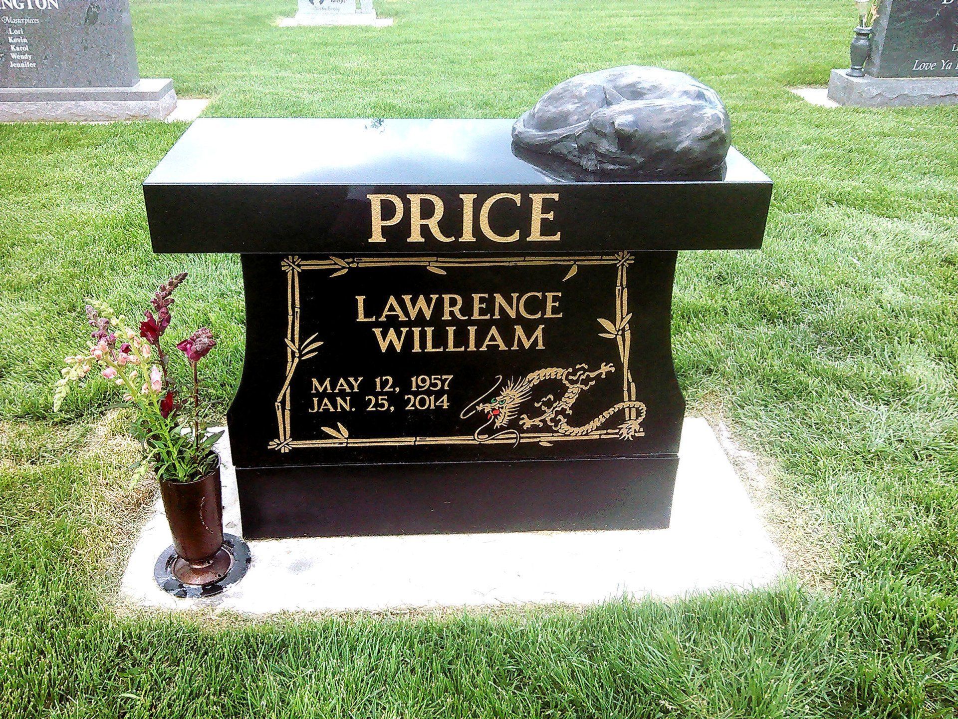 Black gravestone with gold lettering reading PRICE and LAWRENCE WILLIAM, with flowers beside it in a cemetery.