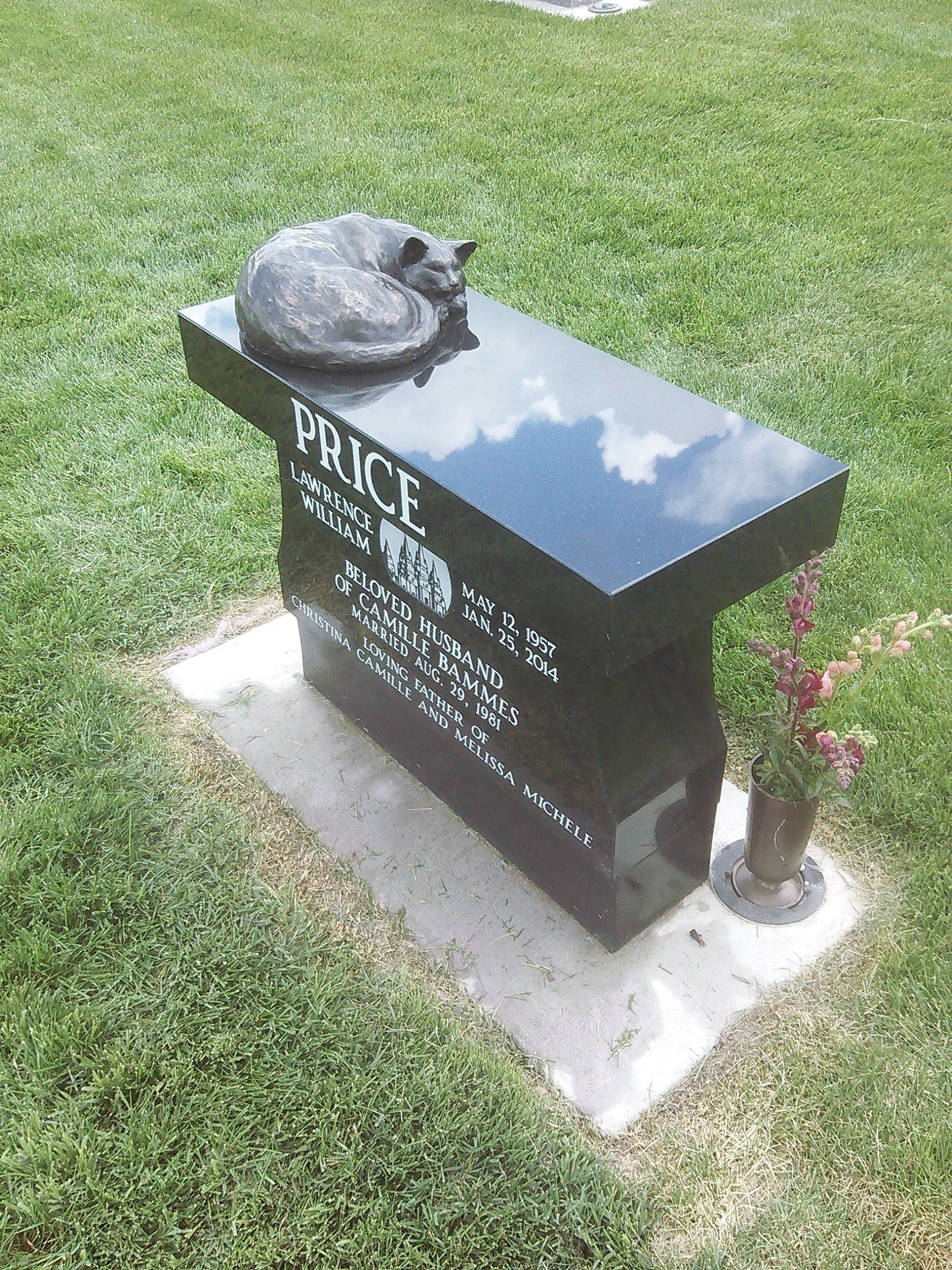 Black memorial bench with engraved plaque and flowers on a grassy lawn.