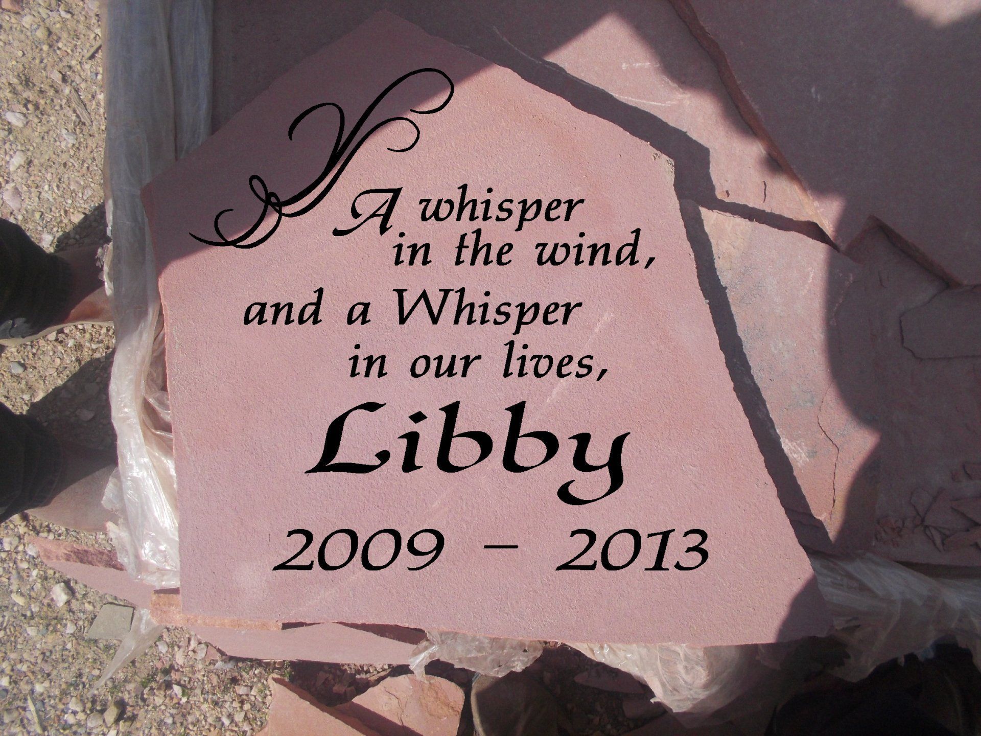 Pink memorial stone engraved “A whisper in the wind…” with “Libby 2009–2013” on a rocky ground