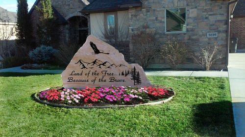 Stone memorial sign in a landscaped flower bed beside a building, with text about the “Land of the Free”.