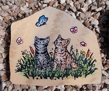 Two kittens in grass with flowers and butterflies on a painted stone.