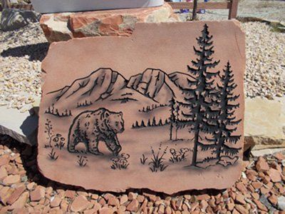 Engraved stone plaque with a bear, pine trees, and mountain landscape in a rocky outdoor setting