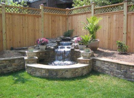 Backyard stone waterfall fountain surrounded by wooden fence, potted plants, and green lawn