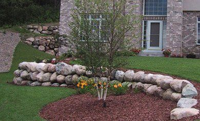 Landscaped front yard with rock border, mulch bed, flowers, and a small tree beside a brick house