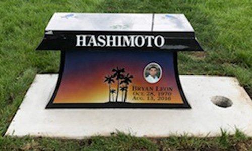 Memorial plaque with “HASHIMOTO” in white on black, set on a concrete base in grass.