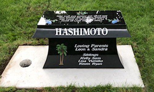 Black gravestone and white base with “HASHIMOTO,” surrounded by grass and flowers.