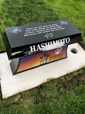 Black memorial plaque with white lettering on a grave marker on grass, with a colorful sunset panel below.