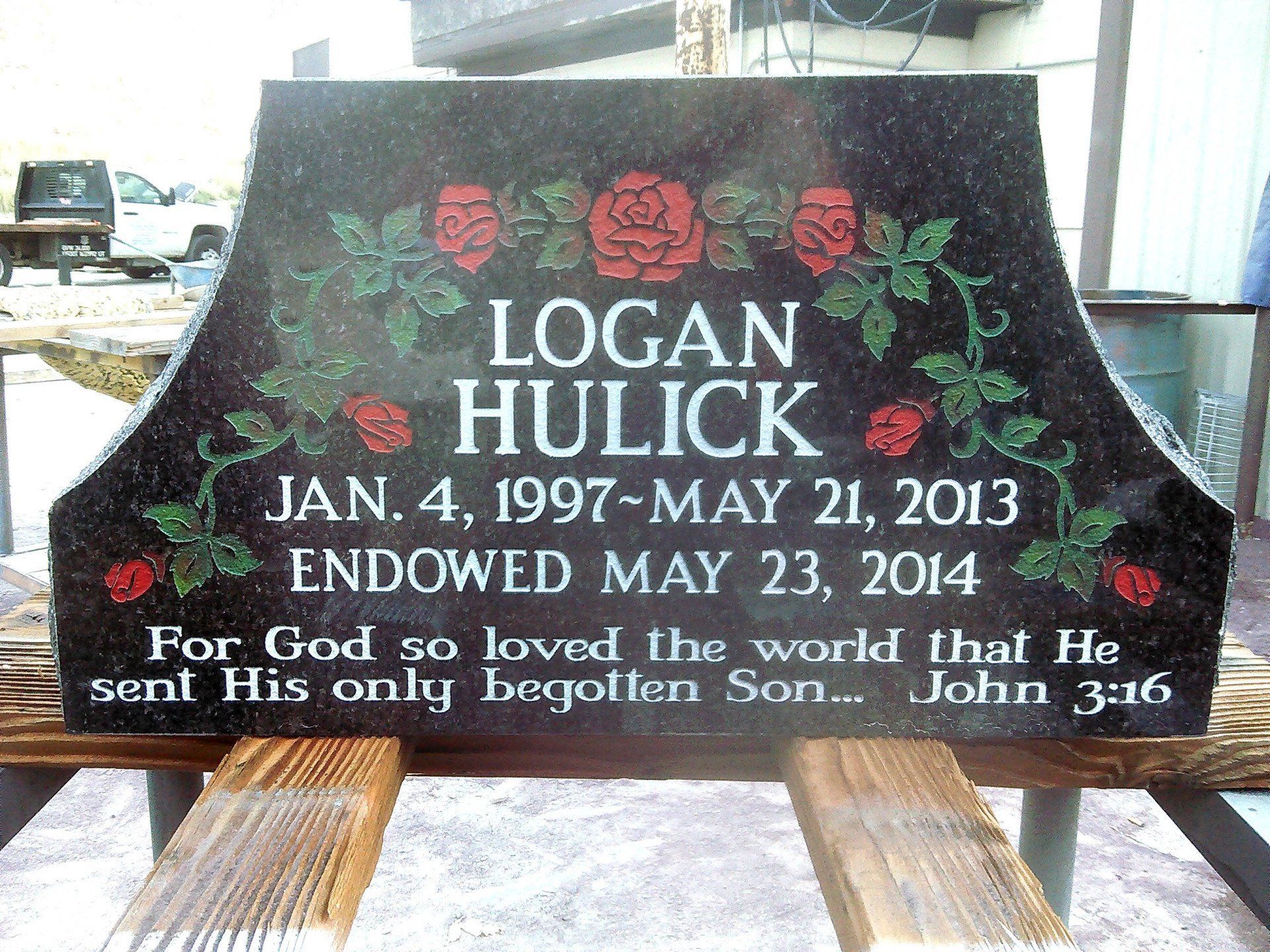 Black granite memorial headstone with red roses reading “Logan Hulick” and Bible verse John 3:16.