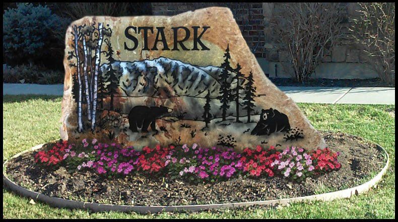 “STARK” memorial sign with painted pine trees, a black bear, and pink flowers in a landscaped garden