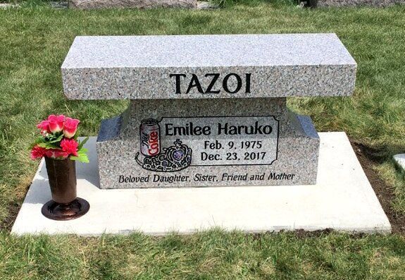 Gravestone reading “TAZOI” with flowers beside a memorial plaque on a grassy lawn.