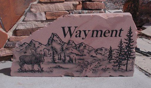 Engraved stone sign reading “Wayment” with mountain, moose, trees, and a log cabin scene