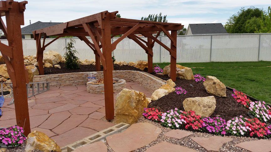 Wooden pergola over a stone path beside a flower bed with pink blooms and large rocks in a landscaped yard