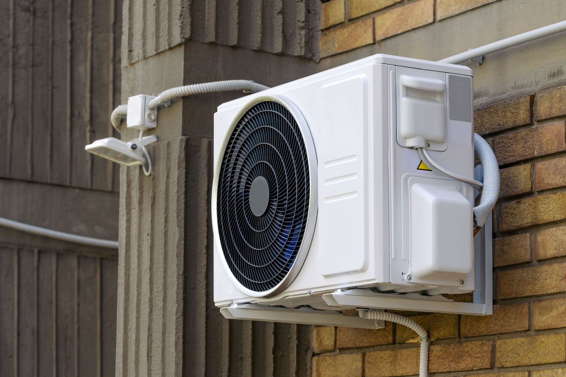 A white outdoor air conditioning condenser unit mounted on a brick wall beside a concrete pillar.