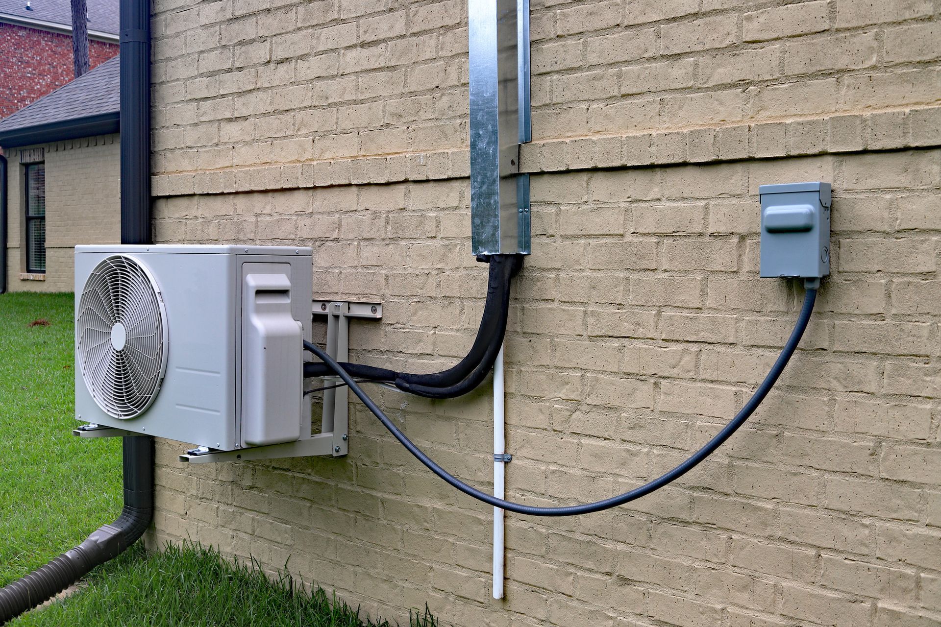 An outdoor mini-split AC unit mounted on a brick wall, connected to a metal conduit and a grey electrical disconnect box.