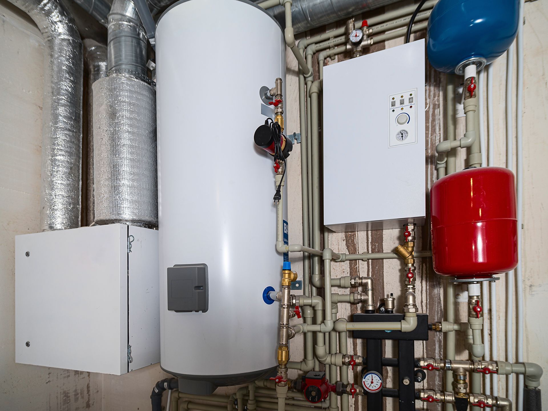 A white water heater tank, heating unit, control box, and red and blue expansion tanks with pipes in a utility room.