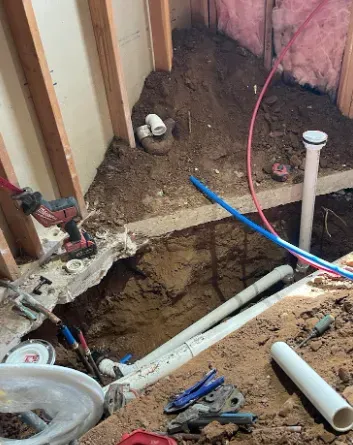 Plumbing construction inside a home, showing an open trench in the dirt floor, exposed pipes, and stud wall framing.