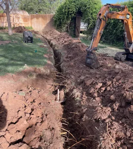 A bright orange excavator sits in a grassy yard next to a long, deep trench dug into the reddish-brown soil.