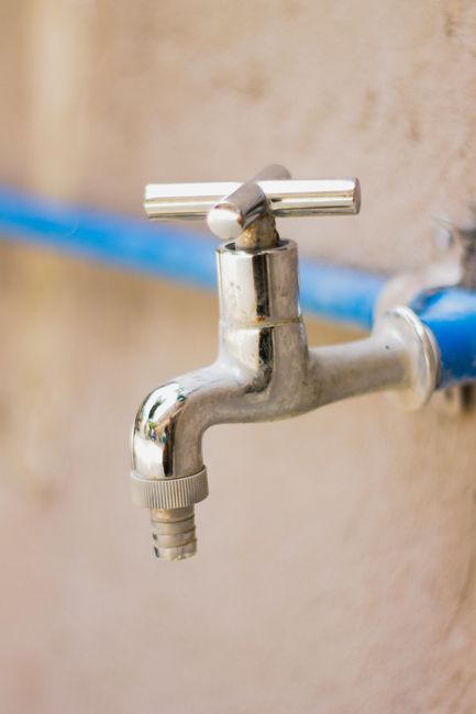 A silver metal water spigot attached to a blue pipe against a beige wall.