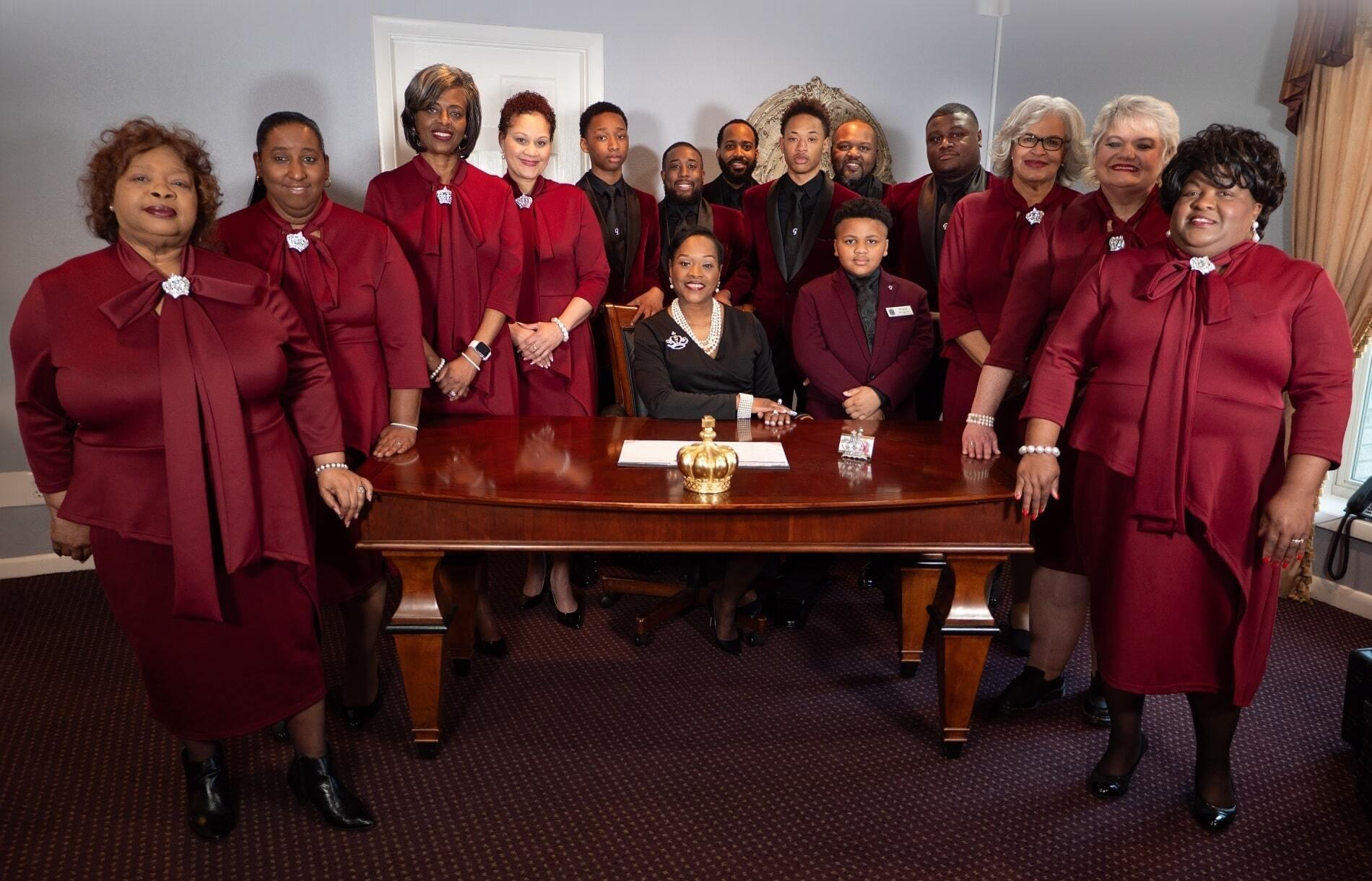 Group in matching burgundy outfits gathered around a desk in an office, with one person seated at the center.