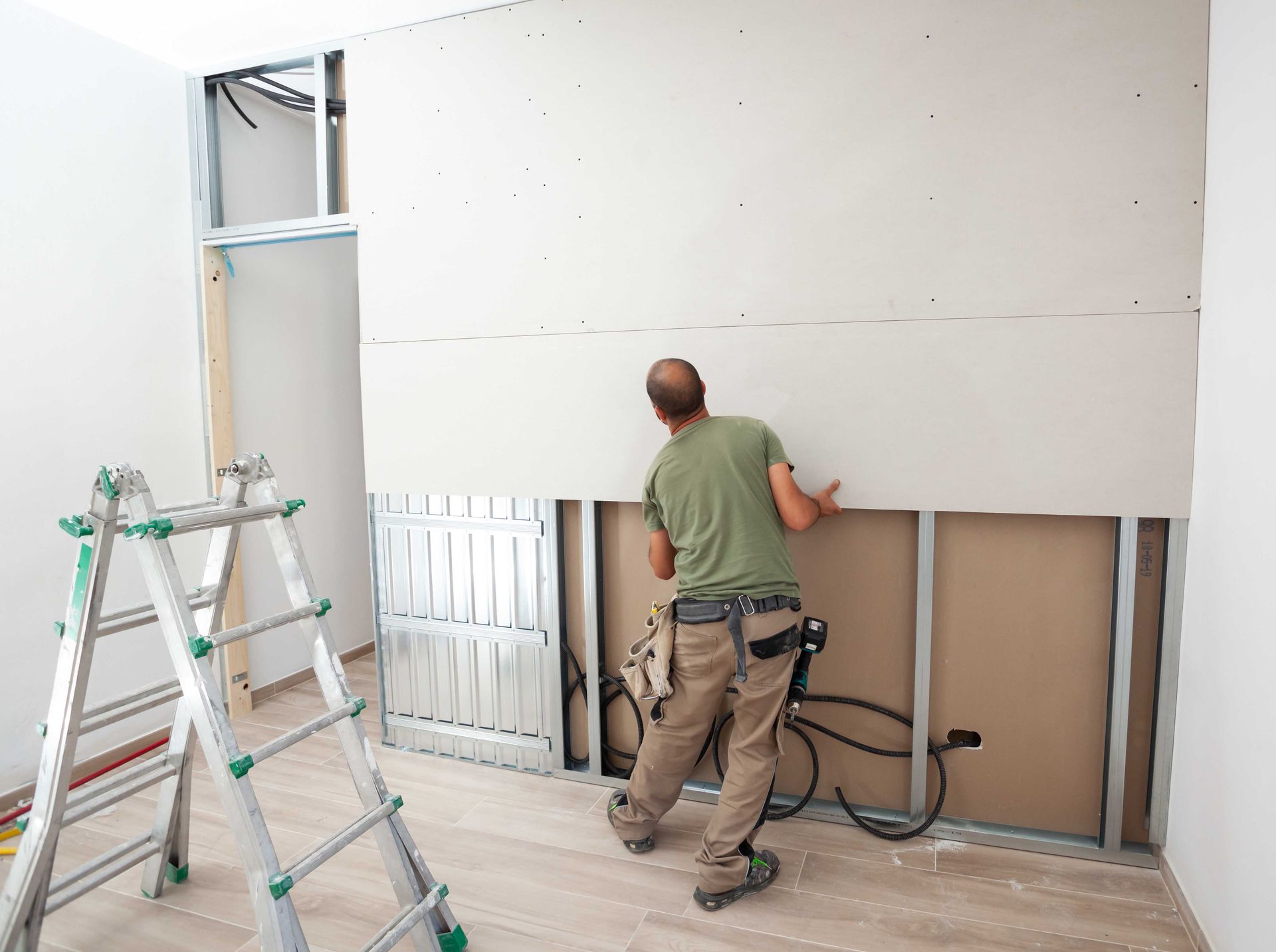 Un ouvrier installe des plaques de plâtre dans une pièce où se trouvent des échelles et des outils de construction.