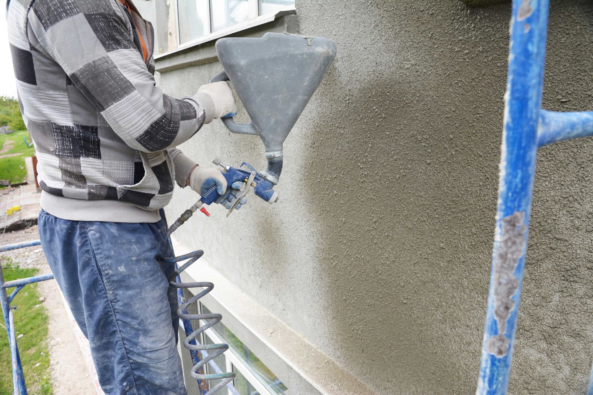 Un ouvrier projette du stuc sur un mur extérieur à côté d'un échafaudage bleu.