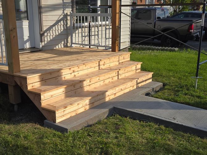 Des marches en bois mènent à un porche jouxtant une maison, avec une cour gazonnée et une voiture garée en arrière-plan.