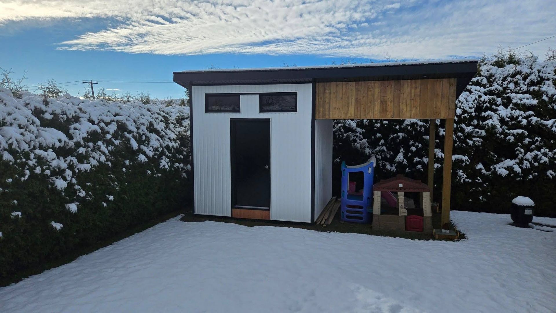 Cour arrière enneigée avec un petit abri de jardin et une terrasse couverte sous un ciel nuageux