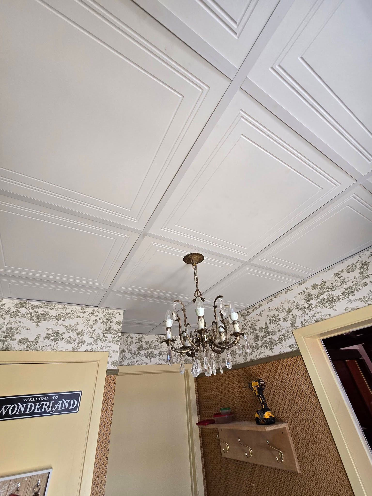 Plafond à panneaux blancs avec moulures décoratives, un lustre, du papier peint à motifs floraux et une étagère murale dans une pièce