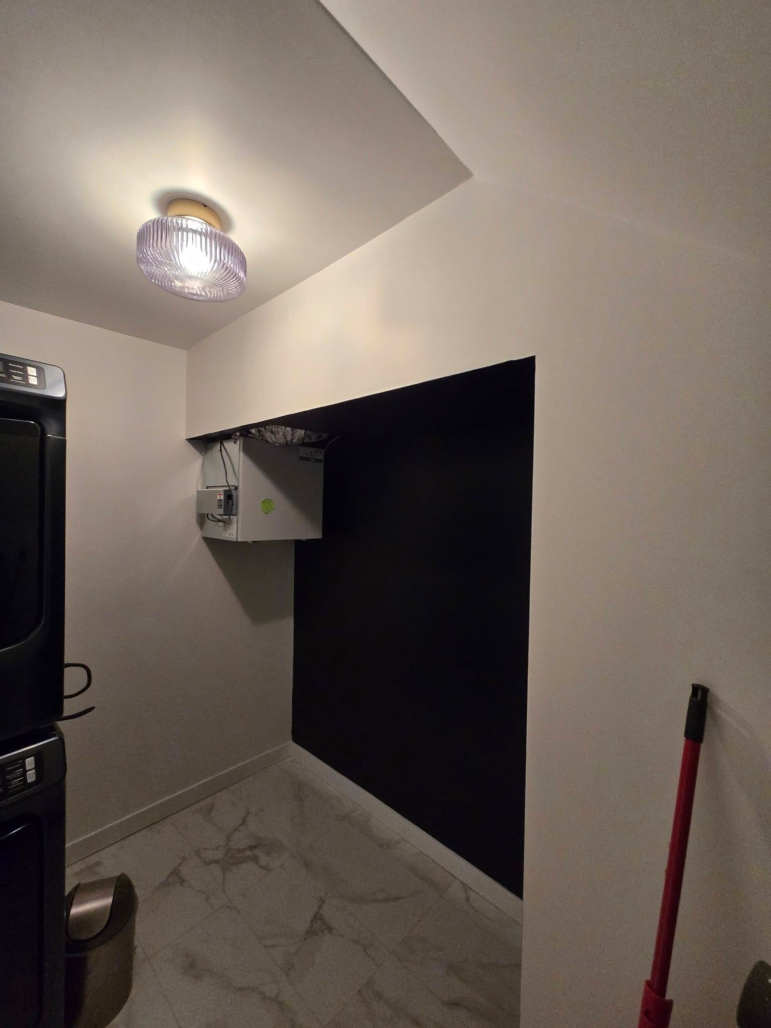 Petite buanderie avec machine à laver, ouverture dans le mur noir, plafonnier et balai rouge appuyé sur la droite.