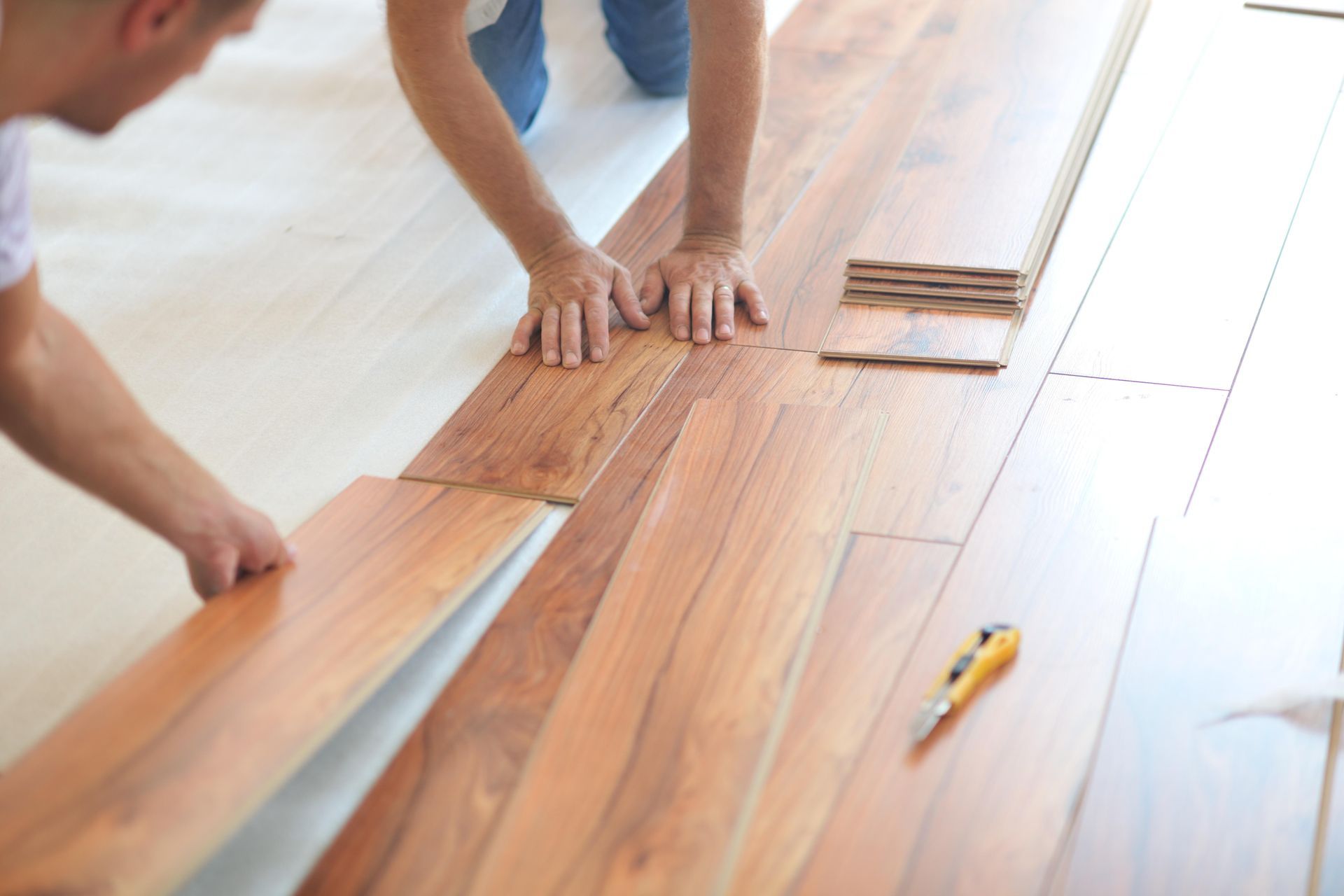 Des mains installent des lames de parquet avec une truelle et un couteau utilitaire à proximité.