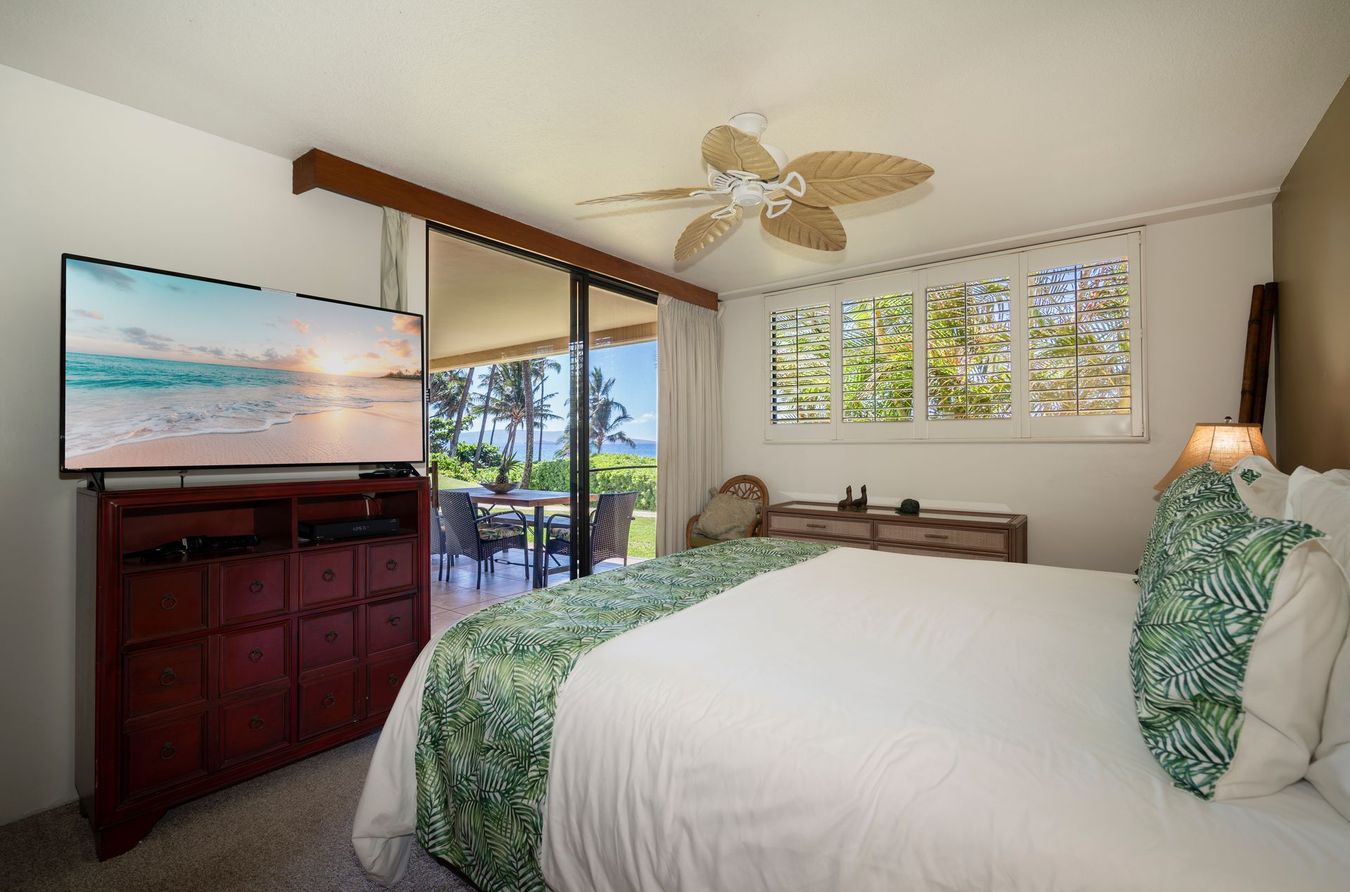 Bedroom with bed, TV, and sliding glass door to a patio with a view of greenery and the ocean.