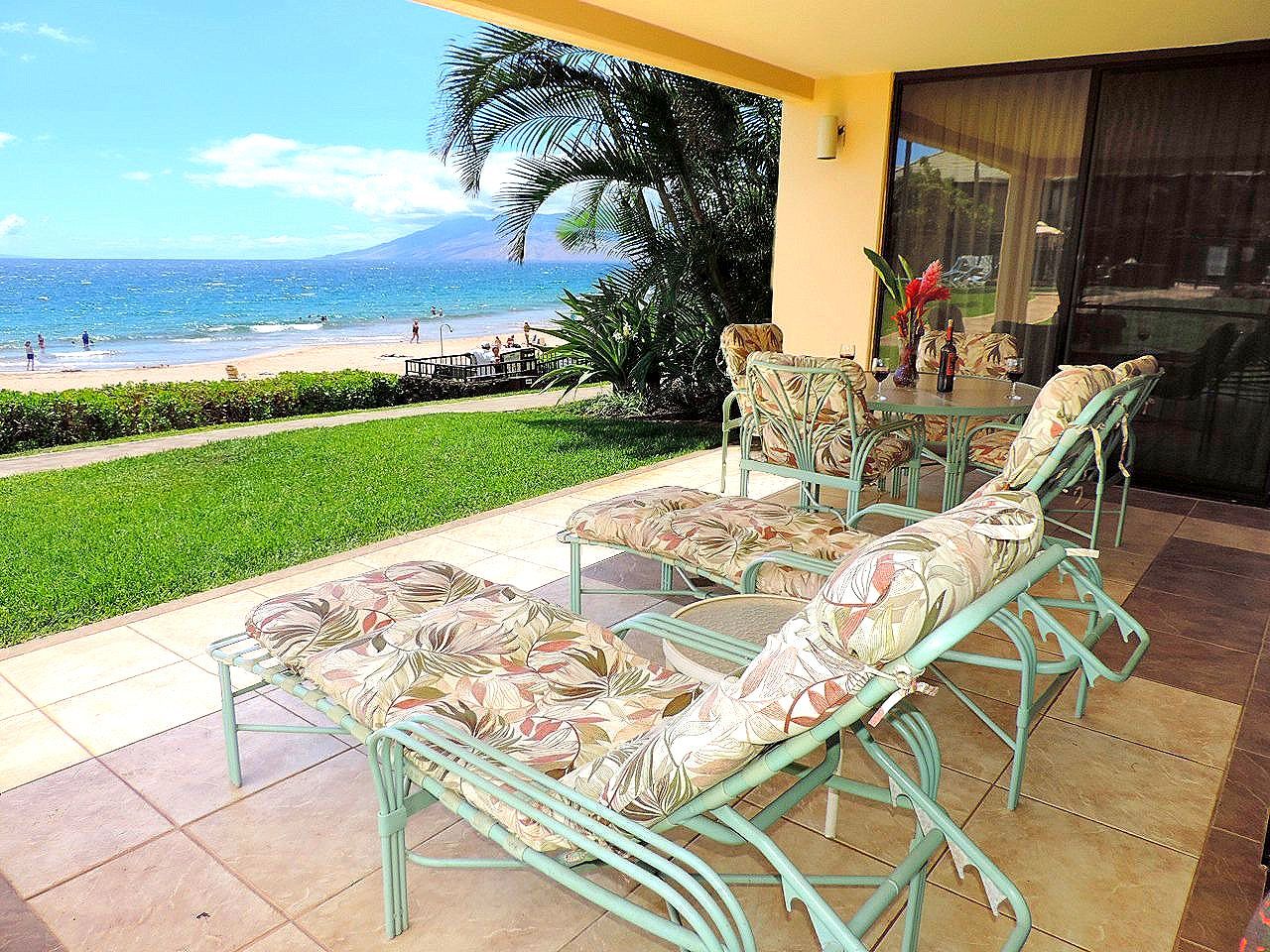 Patio overlooking beach, with lounge chairs and table. Ocean and grass in the background. Sunny day.