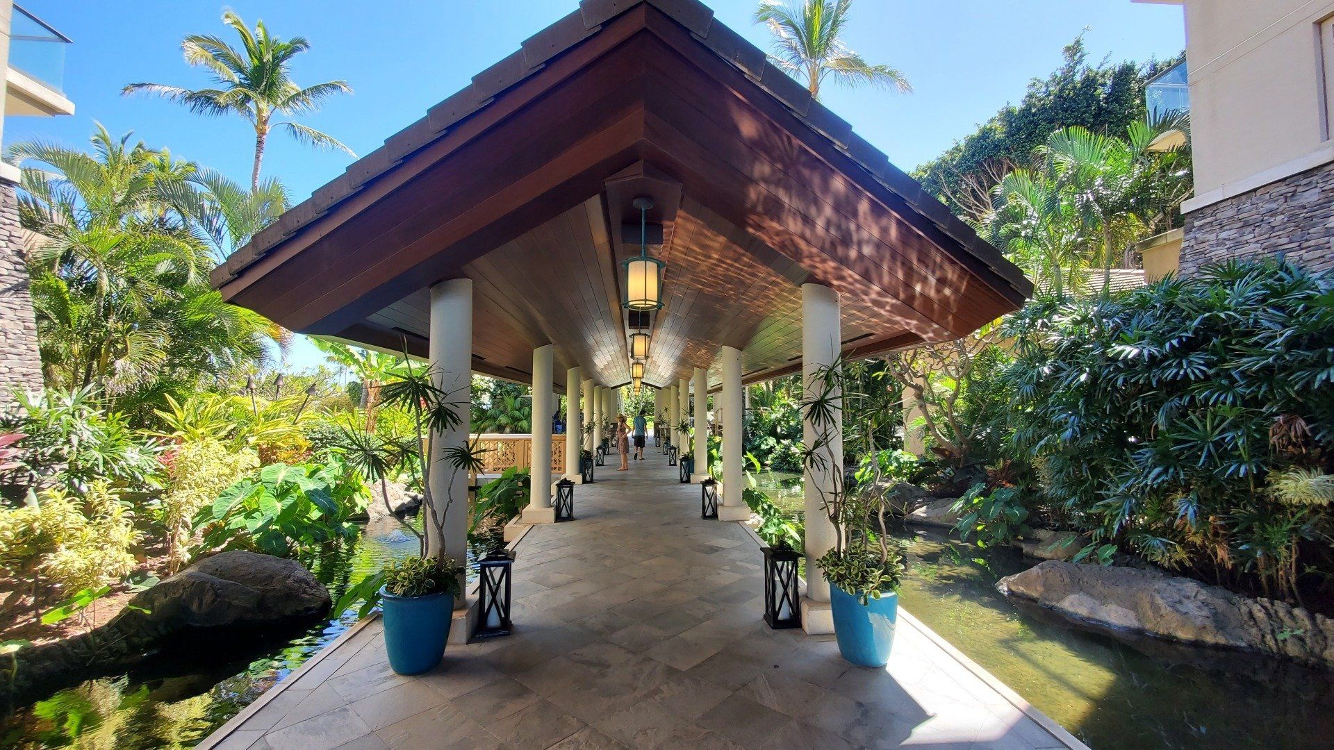 Covered walkway with hanging lights, flanked by lush greenery and water features.