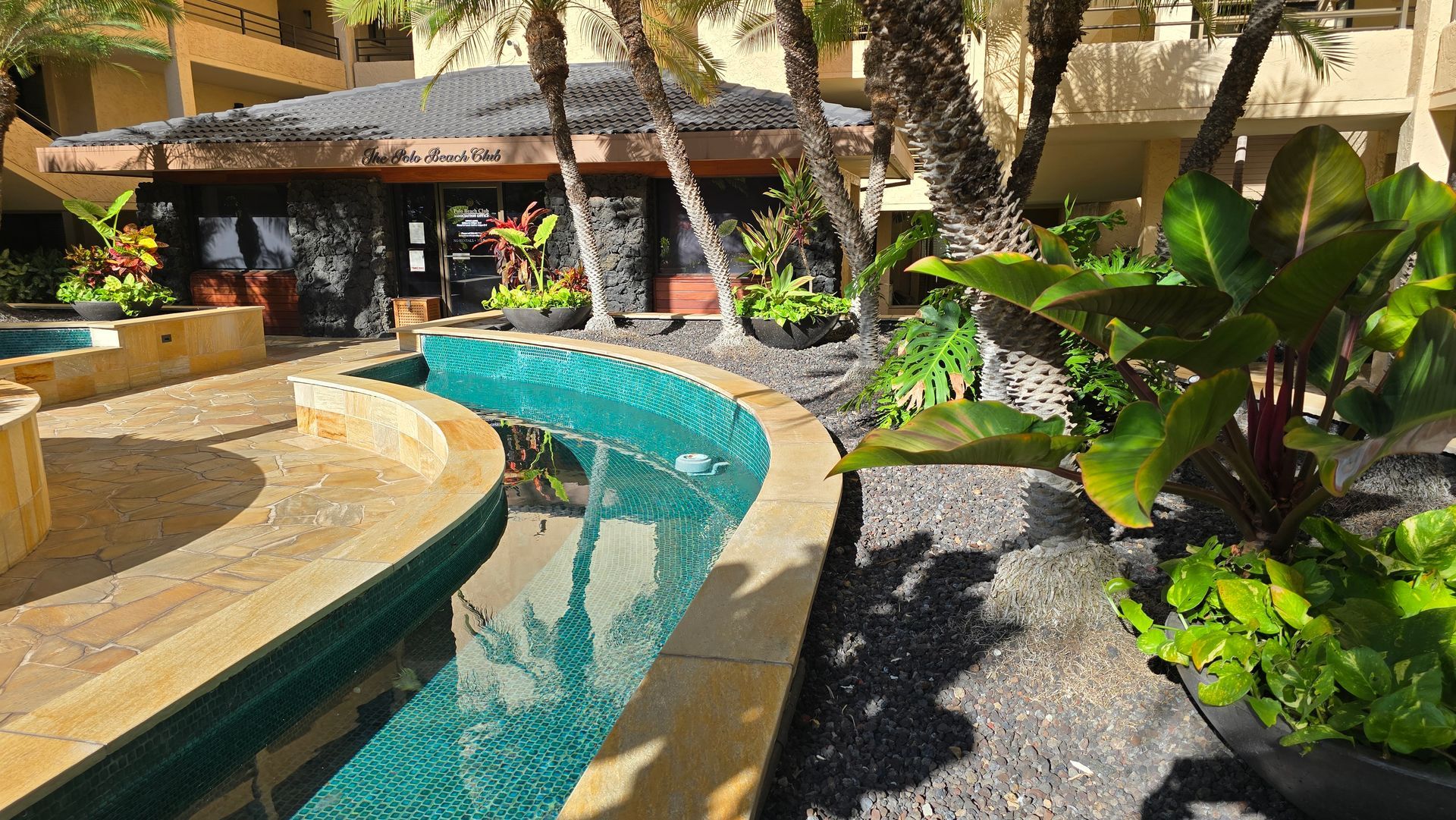 Pool area with turquoise water channel, lush plants, and a building with a dark roof.