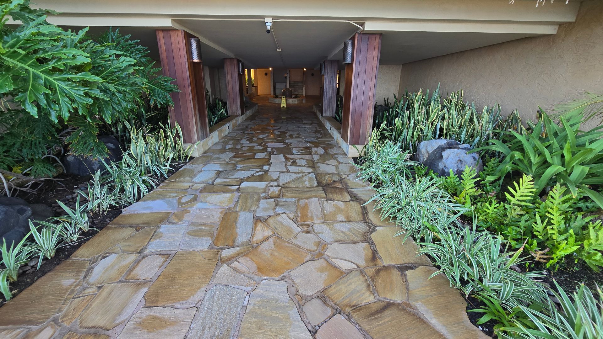 Stone pathway leading through a covered walkway, flanked by lush green plants and stone columns.