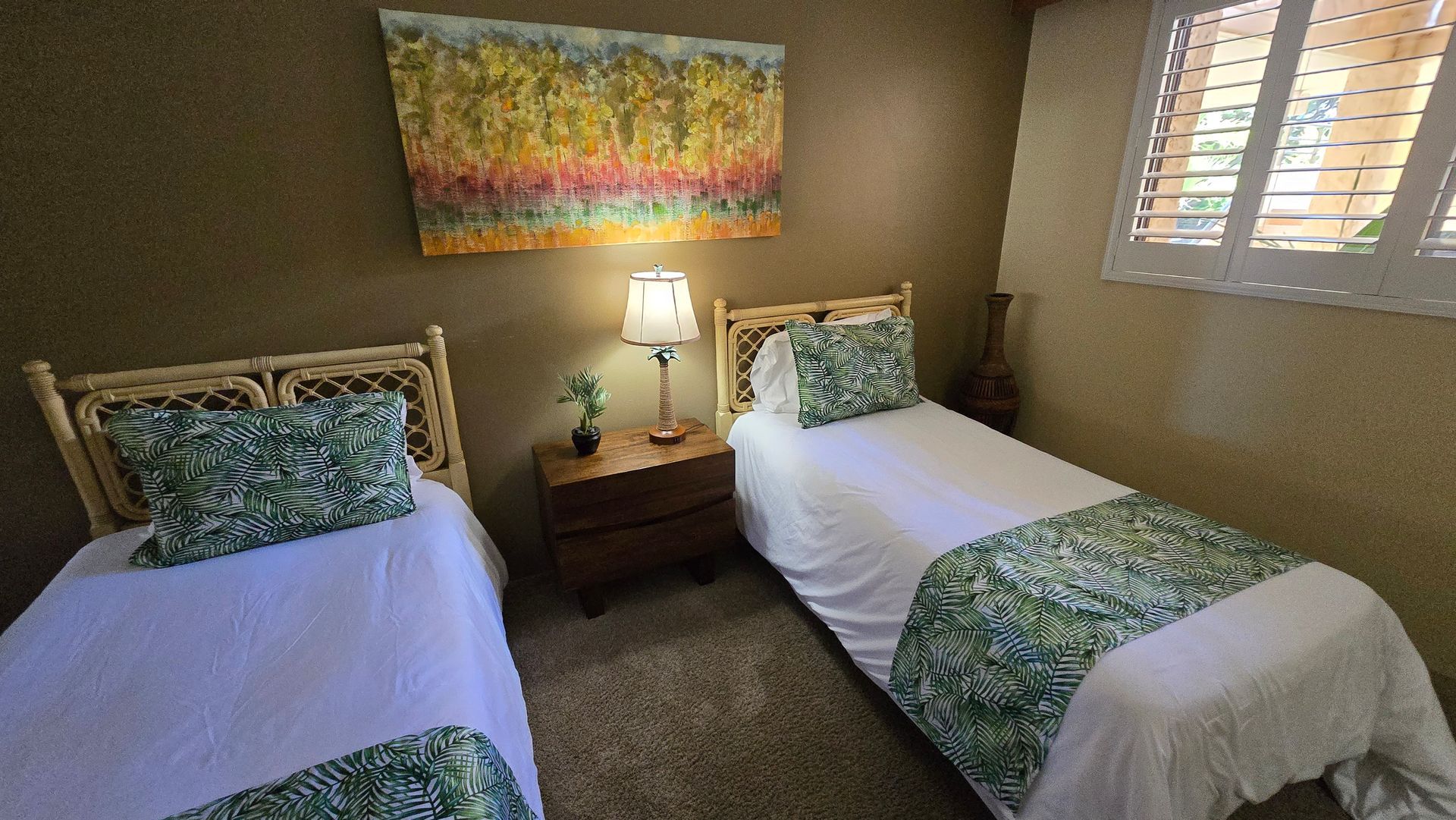 Two twin beds in a bedroom with a nightstand between them, art on the wall, and a window with shutters.